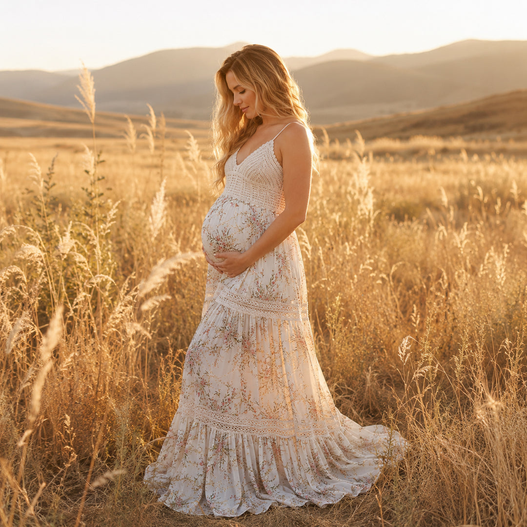 A pregnant woman stands in profile in a field of dry tall grass with mountains in the background, wearing an apricot bottom floral print maternity maxi dress with spaghetti straps, a crochet bodice, and lace-trimmed tiered panels on the skirt that pools on the ground.